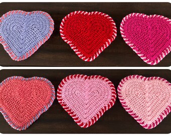 May include: Six crocheted hearts in various shades of pink, red, and purple. The hearts are arranged in two rows of three on a dark wood surface.