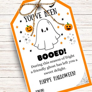 May include: A Halloween-themed gift tag with an orange border and a string bow. The tag features a friendly ghost illustration, pumpkins, stars, and the text "YOU'VE BEEN BOOED!" and "HAPPY HALLOWEEN!" on a white background.
