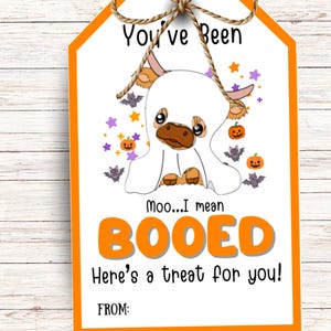 May include: An orange Halloween gift tag featuring a cartoon cow in a ghost costume. Text on the tag reads "You've Been BOOED! Here's a treat for you!" with a space for "FROM:". The tag is decorated with bats, pumpkins, and stars, and tied with twine.