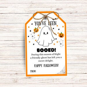 May include: A Halloween-themed gift tag with an orange border and a jute twine bow. The tag features a cartoon ghost, pumpkins, stars, and the text "YOU'VE BEEN BOOED!" and "HAPPY HALLOWEEN!" on a white background.