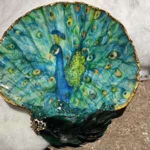 Op de afbeelding: Een decoratieve schelpschaal met een pauwenontwerp. De pauw is afgebeeld met blauwe, groene en gele veren. De schaal heeft een goudkleurige rand en is versierd met zwarte veren en een juwelen accent.