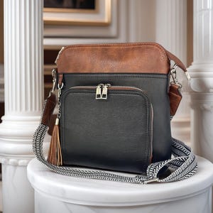 May include: A two-tone crossbody bag featuring a black main body and a brown top panel. The bag includes a front zippered pocket, silver-tone hardware, a brown leather-like tassel charm, and a wide, black and white patterned fabric shoulder strap.