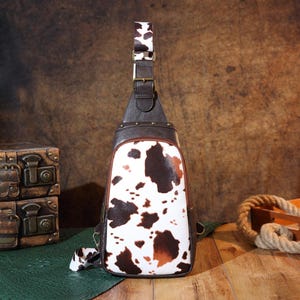 May include: A brown and white cowhide-patterned sling bag with a brown leather strap and accents. The bag has a zippered front pocket and a brown leather strap. The bag is made of leather and has a unique, eye-catching design.
