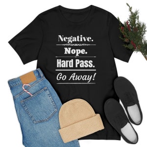 Op de afbeelding: Zwart T-shirt met de witte tekst "Negative. Nope. Hard Pass. Go Away!". Het shirt wordt gecombineerd met een blauwe jeans, een beige muts en zwarte schoenen. Een zuurstok zit in de jeanszak.