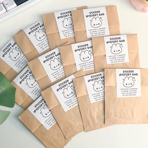 May include: A collection of brown paper bags labeled "Sticker Mystery Bag" with a cute bunny graphic. Each bag contains a white label with the bag's name and contents. The bags are arranged on a white surface.