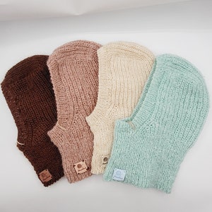May include: Four knitted balaclavas in shades of brown, pink, cream, and mint green are displayed. Each balaclava has a textured knit pattern and a small, rectangular tag. The balaclavas are arranged on a white surface.