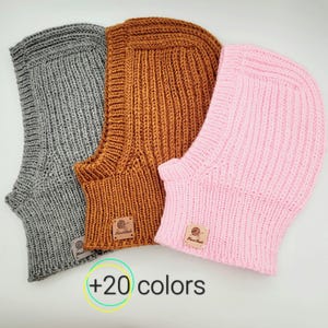 May include: Three knitted balaclava hats in gray, brown, and pink. Each hat has a small leather tag with the word "Handmade" on it. The text " +20 colors" is visible below the hats.