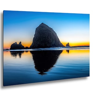May include: A scenic photograph of a coastal landscape featuring a large rock formation silhouetted against a vibrant sunset. The sky transitions from blue to orange and yellow, reflected in the wet sand and water.