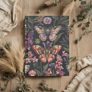 May include: A hardcover journal with a dark background featuring a floral and butterfly design. The cover showcases two large moths with intricate wing patterns, surrounded by pink and purple flowers. The journal is rectangular and lies on a wooden surface.