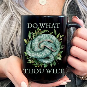 May include: Black ceramic mug featuring a coiled turquoise snake illustration surrounded by green leaves. The mug has the text "DO WHAT THOU WILT" in white lettering. The mug is held by a person.