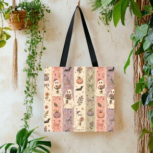 May include: A tote bag with black shoulder straps featuring a repeating vertical pattern of ghosts holding flower bouquets, pumpkins, bats, bows, and stars. The background consists of alternating stripes in shades of cream, pink, and purple.