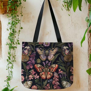 May include: Black tote bag featuring a vibrant butterfly and floral pattern. The design includes various butterfly species in shades of brown, purple, and red, set against a dark background with pink and purple flowers. The bag has black handles.