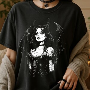 May include: A black short-sleeved t-shirt featuring a white graphic print of a winged figure with horns, wearing a corset-style top. The illustration displays a detailed character with dark hair and wings, centered on the front of the garment.