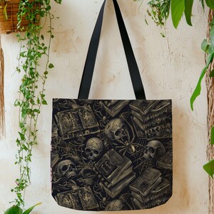 May include: Black tote bag with a repeating pattern of skulls, books, and spiderwebs in shades of black and gold. The bag has black handles and is hanging against a neutral background. The design gives a gothic or Halloween theme.