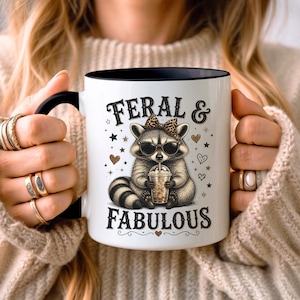May include: A white ceramic mug with a black handle and rim, featuring a graphic of a raccoon wearing sunglasses and a leopard print bow, holding a plastic cup with a straw. The text FERAL & FABULOUS is printed in black above and below the raccoon.