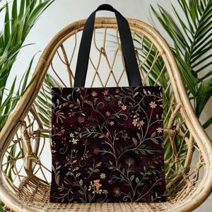 May include: A tote bag with a dark burgundy background and a floral pattern in gold, green, and pink. The bag has black handles and is displayed on a wicker chair.