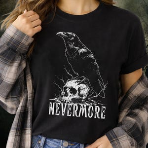 May include: Black t-shirt featuring a white graphic of a raven perched atop a skull, with the word "NEVERMORE" in a bold, gothic font below. The design is a detailed illustration, perfect for fans of dark aesthetics.