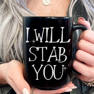 May include: Black ceramic mug with the text "I WILL STAB YOU" in white, with star accents. The mug has a large handle and is held by a person. The mug is a standard size for coffee or tea.