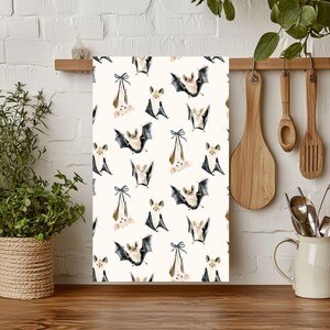 May include: A cream-colored kitchen towel with a repeating pattern of bats in various poses. The bats have black wings, tan bodies, and some are hanging upside down with blue bows. The towel is hanging on a wooden rack.