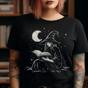May include: Black t-shirt featuring a detailed illustration of a witch reading a book under a crescent moon and stars. The witch is wearing a pointed hat and robe. The design is in white against the black fabric.
