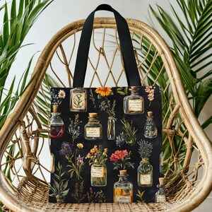 May include: Black tote bag featuring a botanical illustration of various glass bottles filled with colorful liquids and flowers. The bottles are labeled with handwritten text. The bag has black handles and is set against a wicker chair and leafy green plants.