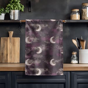 May include: A dark purple kitchen towel with a celestial design. The towel features white crescent moons, clouds, and stars. The background has a mix of purple and gray hues, creating a night sky effect.