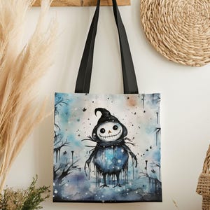 May include: A black tote bag with black handles. The bag features a watercolor illustration of a spooky scarecrow character with a smiling face, wearing a black hat and scarf, set against a blue and white background.
