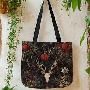 May include: A black tote bag with a festive design featuring red ornaments, berries, and a deer skull with antlers. The bag has black handles and is hanging against a textured wall with greenery.
