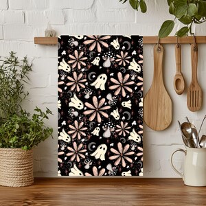 May include: A rectangular kitchen towel featuring a repeating pattern on a black background. The design includes pale pink flowers, cream-colored ghosts, grey crescent moons, small mushrooms, and tiny starbursts scattered across the fabric surface.