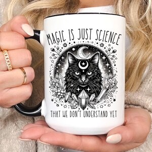 May include: White ceramic mug with a black handle and rim. The mug features a black cat illustration with a crescent moon and crystal accents, and the text "MAGIC IS JUST SCIENCE THAT WE DON'T UNDERSTAND YET."