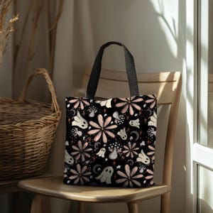 May include: A black tote bag featuring a repeating pattern of light pink flowers, white ghosts, spotted mushrooms, and small crescent moons. The bag has two black fabric handles and a rectangular shape with a flat bottom and a printed design.