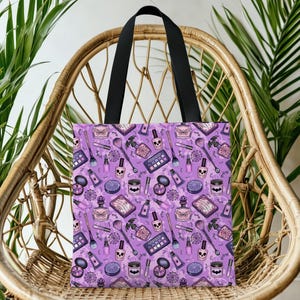 May include: A purple tote bag with black handles, featuring a repeating pattern of makeup items like eyeshadow palettes, nail polish, and perfume bottles. The design includes skull and bat motifs.