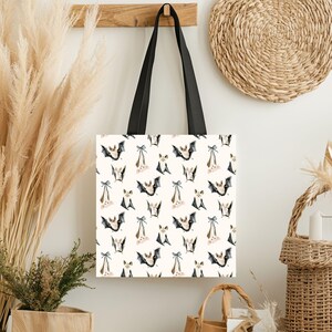 May include: A white tote bag with black shoulder straps featuring a repeating pattern of watercolor-style bats in various poses, including some hanging upside down with small blue bows tied to their feet against a light cream background.
