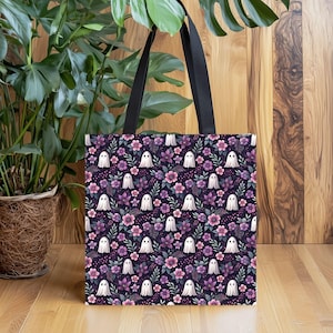 May include: A black tote bag featuring a repeating pattern of small white ghosts interspersed with purple and pink flowers and green foliage. The bag has two black fabric shoulder straps attached to the top edge of the rectangular body.