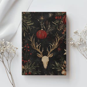 May include: A rectangular journal with a dark background featuring a deer skull, antlers, red ornaments, and botanical elements. The cover has a festive, vintage aesthetic with a Christmas theme. The design includes berries and foliage.
