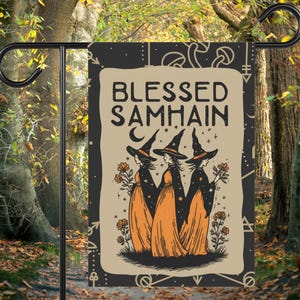 May include: A garden flag featuring the text "Blessed Samhain" with an illustration of three witches in black hats and orange dresses. The flag is mounted on a black metal stand, set against a backdrop of trees.