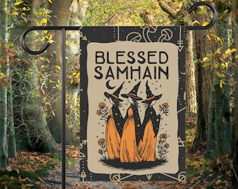 Könnte beinhalten: Eine Gartenflagge mit dem Text "Blessed Samhain" und einer Illustration von drei Hexen in schwarzen Hüten und orangefarbenen Kleidern. Die Flagge ist auf einem schwarzen Metallständer vor Bäumen befestigt.