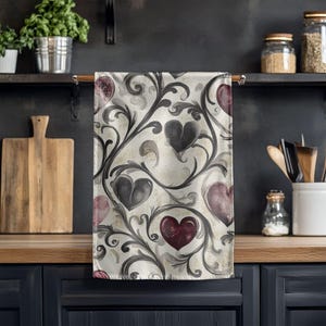 May include: A kitchen towel with a repeating pattern of hearts and swirling vines. The hearts are in shades of burgundy, gray, and black, set against a cream-colored background. The towel is hanging on a wooden rod.