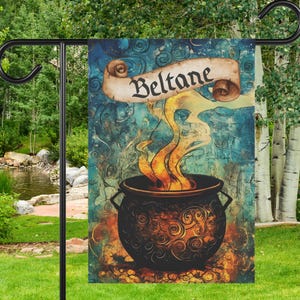 May include: A garden flag featuring a cauldron with flames and steam rising from it. The flag has a blue and teal background with the word "Beltane" on a scroll. The flag is hanging on a black metal garden stake.