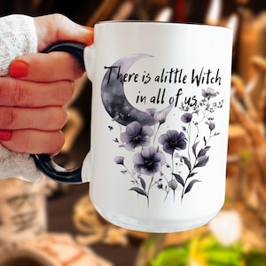 May include: White ceramic mug with a black handle and rim. The mug features a crescent moon and floral design in shades of purple and gray, with the text "There is a little Witch in all of us."