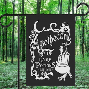 May include: Black garden flag with white text that reads "Apothecary Rare Potions - EST 1692." The flag features a mortar and pestle, a bottle, and a lantern design. The flag is displayed on a black metal stand in a forest setting.