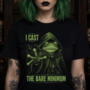 May include: A black t-shirt featuring a green graphic of a toad wearing a hooded cloak and casting a spell with its hand. The text above the toad reads I CAST and the text below reads THE BARE MINIMUM in a bold, sans-serif green font.