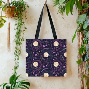 May include: A square tote bag with black shoulder straps featuring a repeating pattern of full moons, purple tulip-like flowers, and dark green leafy vines against a deep navy blue background. The design includes small white star accents.