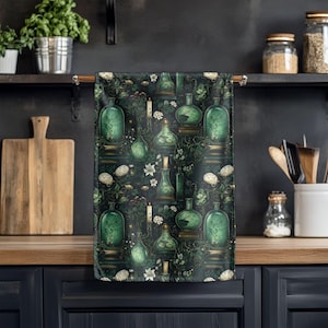 May include: A dark green kitchen towel with a repeating pattern of green potion bottles, candles, and white flowers. The towel is hanging on a wooden rod in a kitchen setting, with a wooden cutting board and jars in the background.