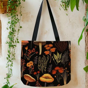 May include: Black tote bag featuring a colorful mushroom and floral pattern. The design includes orange, yellow, and brown mushrooms and flowers on a black background. The bag has black straps.