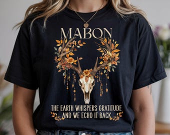 May include: Black t-shirt featuring a deer skull design with the word "MABON" above it. The design includes a floral wreath with fall colors and the text "THE EARTH WHISPERS GRATITUDE AND WE ECHO IT BACK".