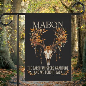May include: Black garden flag featuring a deer skull with antlers adorned with autumn leaves and flowers. The word "MABON" is at the top, with the text "THE EARTH WHISPERS GRATITUDE AND WE ECHO IT BACK" below the skull.