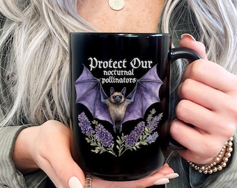 May include: A black ceramic mug featuring a graphic of a brown bat with purple wings spread wide, positioned above purple lavender flowers. The text "Protect Our nocturnal pollinators" is printed in white above the bat illustration.