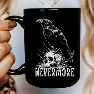 May include: Black ceramic mug featuring a white illustration of a raven perched on a skull. The word "NEVERMORE" is printed in white below the skull. The mug has a black handle.
