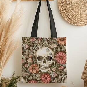 May include: A tote bag featuring a skull design surrounded by flowers and mushrooms in shades of brown, pink, and green. The bag has black handles and is made of a durable fabric, perfect for carrying groceries or books.